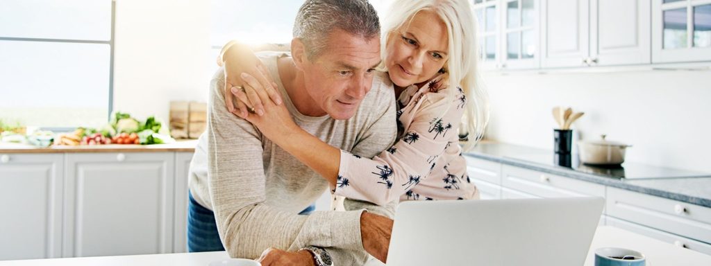 Retired couple reviewing their monthly retirement budget and bills at a kitchen table with a laptop